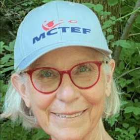 Smiling elderly woman wearing a light blue cap and red glasses outdoors.
